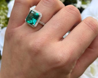 Könnte beinhalten: Ein silberner Ring mit einem großen smaragdgrünen Edelstein, der von kleineren klaren Edelsteinen umgeben ist.