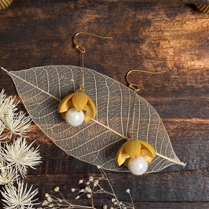 May include: A pair of gold earrings with a delicate design featuring a small, dried flower with a pearl in the center. The earrings are displayed on a dried leaf with a wooden background.