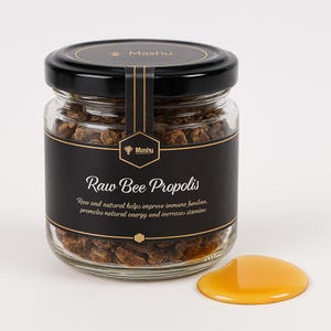 Puede incluir: Un tarro de vidrio lleno de propóleo de abeja crudo, un producto natural que ayuda a mejorar la función inmunitaria, promueve la energía natural y aumenta la resistencia. El tarro tiene una etiqueta negra con el texto "Propoleo de abeja crudo" y el nombre de la marca "Mashu". Una pequeña cantidad de miel se derrama sobre la superficie blanca.