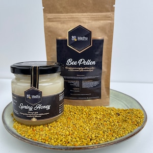 May include: A glass jar of light-colored honey with a black label that reads "Madhu Bee Products Spring Honey Creamy & Light It's produced from nectar and wild flowers from meadows of Lithuania" and a brown paper bag with a black label that reads "Madhu Bee Products Bee Pollen Energize your everyday with bee pollen - a clean source of bee-sourced fuel" and a plate of yellow bee pollen.