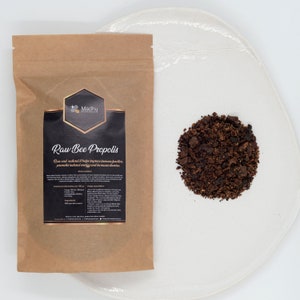 Op de afbeelding: Een bruine papieren zak met een label waarop "Madhu Naturals Raw Bee Propolis" staat. Het label beschrijft het product als rauw en natuurlijk, helpt de immuunfunctie te verbeteren, bevordert natuurlijke energie en verhoogt het uithoudingsvermogen. Een kleine stapel donkerbruine bijenpropolis ligt op een wit bord.