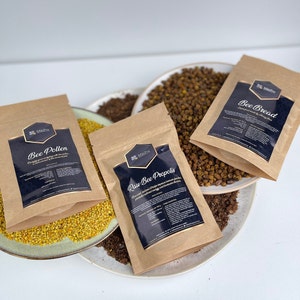 Könnte beinhalten: Vier braune Papiertüten mit schwarzem Text und einem Bienenlogo. Die Tüten enthalten Bienenpollen, rohe Bienenpropolis und Bienenbrot. Die Tüten sind auf einer weißen Oberfläche mit einem Teller gelben Bienenpollens im Vordergrund angeordnet.