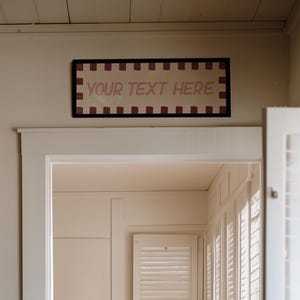 May include: A rectangular wall sign with a dark frame and a white background featuring a red and white checkered border and the text YOUR TEXT HERE in the centre, positioned above a doorway leading into a bedroom with a bed and a potted plant.