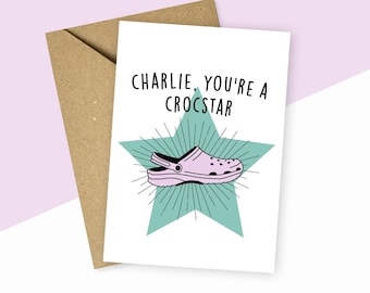 Könnte beinhalten: Eine weiße Karte mit einem hellblauen Stern und einem rosa und schwarzen Croc-Schuh. Der Text auf der Karte lautet "Charlie, du bist ein Crocstar".