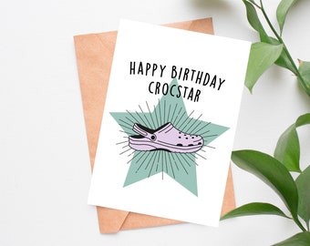 Könnte beinhalten: Eine weiße Karte mit dem Text "Happy Birthday Crocstar" und einer Cartoon-Illustration eines lila und weißen Croc-Schuhs mit einem grünen Stern dahinter.