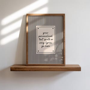 May include: A framed print with a black and white striped background. The print has a rectangular insert with the text "your personalised text, quote or song lyrics go here". The frame is made of wood and sits on a wooden shelf.