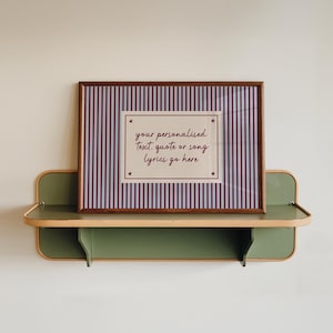 Könnte beinhalten: Ein gerahmter Druck mit einem braunen Holzrahmen, der einen gestreiften Hintergrund in Weiß und Burgunderrot aufweist. Der Druck enthält den Text "your personalised text, quote or song lyrics go here" in handschriftlicher Form. Der Rahmen liegt auf einem grünen Regal.