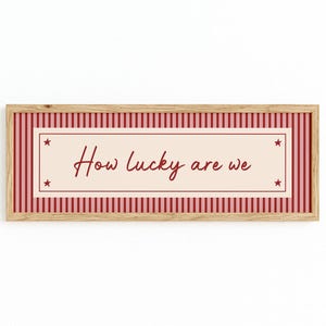 May include: A framed print with the phrase "How lucky are we" in a cursive font. The print features a light beige centre with the text, framed by a pink rectangle with stars. The outer border is pink and white striped, set in a wooden frame.