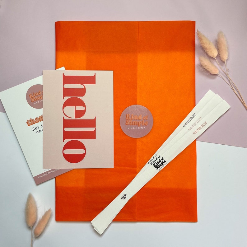 May include: Orange tissue paper with a white card that says "hello" in red letters. The card is next to a white card with the text "Kind of Simple Designs" and the words "Get 1 free next order" on it. There is also a white paper fan with the text "Kind of Simple Designs" on it.
