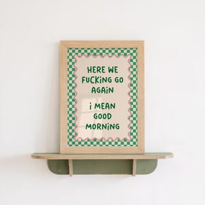 Könnte beinhalten: Ein gerahmter Druck mit dem Text "HERE WE FUCKING GO AGAIN, I MEAN GOOD MORNING" in Grün und Rosa. Der Druck hat einen hellen Holzrahmen und einen grün-weiß karierten Rand. Er steht auf einem grünen Regal.