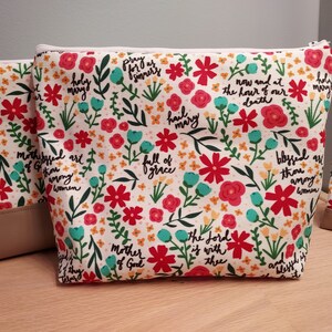 May include: A set of floral print cosmetic bags with a cream-colored background, red and teal flowers, and script text. The bags have a beige bottom and a zipper closure. The text includes phrases like "Hail Mary" and "Mother of God."