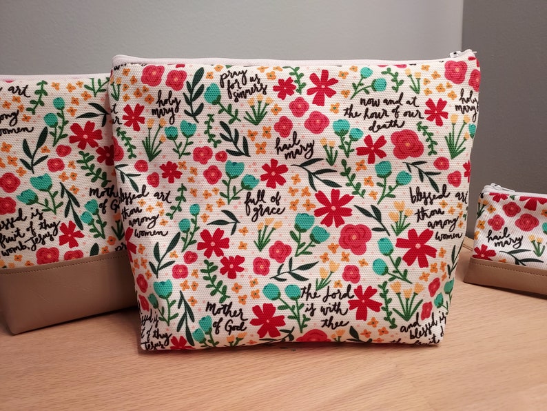 May include: A set of floral print cosmetic bags with a cream-colored background, red and teal flowers, and script text. The bags have a beige bottom and a zipper closure. The text includes phrases like "Hail Mary" and "Mother of God."