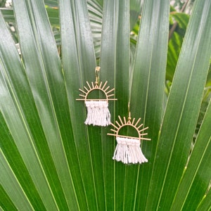 May include: A pair of gold sunburst earrings with white macrame fringe hanging from a green leafy background.