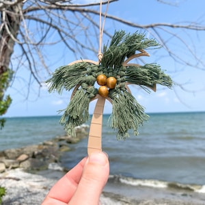 May include: A wooden palm tree ornament with green yarn fringe and brown wooden beads. The ornament is hanging from a string and is being held in a hand against a background of a beach and blue water.