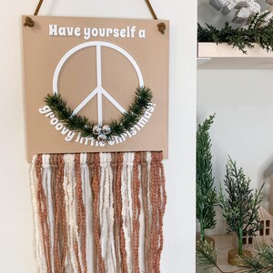 May include: A tan wooden sign with a peace symbol and the words "Have yourself a groovy little Christmas." The sign has a macrame fringe in shades of brown and white. A brown cord is used for hanging.