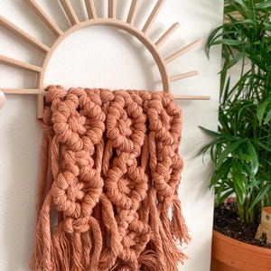 May include: A decorative wall hanging featuring a sunburst design in light brown wood, with a woven macrame piece in a warm terracotta color. The macrame has a textured, knotted pattern with fringe.