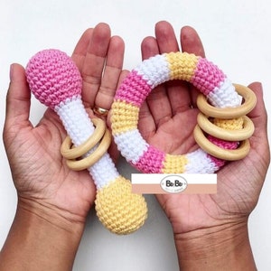 Pode incluir: Dois mordedores de crochê para bebês, um é um anel com listras rosa, brancas e amarelas e o outro é um chocalho com um esquema de cores rosa, branco e amarelo. Ambos os mordedores têm anéis de madeira.
