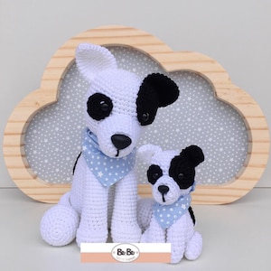Pode incluir: Dois peluches de cachorro de crochê, um maior que o outro, ambos brancos com manchas pretas e bandanas com estampa de estrelas azuis e brancas. Os peluches estão sentados em frente a uma moldura de madeira em forma de nuvem com um fundo azul claro.