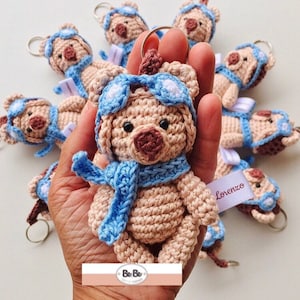 Pode incluir: Uma coleção de chaveiros de urso de peluche de crochê com óculos de aviador azuis e cachecóis. Os ursos são bege com narizes marrons e olhos pretos. Os chaveiros são etiquetados com o nome "Lorenzo".
