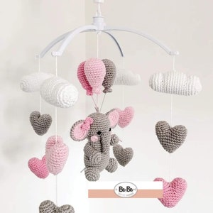 Pode incluir: Um móbile de bebé branco com um elefante de crochet cinzento, corações rosa e cinzentos, nuvens brancas e balões rosa e cinzentos. O móbile tem uma estrutura de metal branco e está pendurado num gancho de teto branco.
