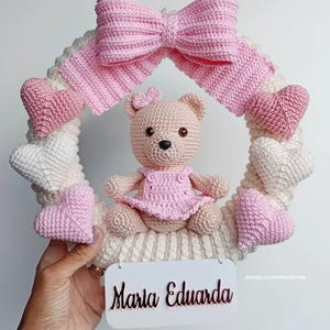 Può includere: Una ghirlanda all'uncinetto fatta a mano con un orsacchiotto marrone chiaro con un vestito rosa, circondato da cuori rosa e bianchi. Un grande fiocco rosa adorna la parte superiore. È attaccato un cartello bianco con il nome "Maria Eduarda".