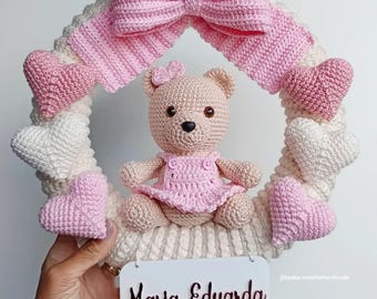 May include: A handmade crochet wreath featuring a light brown teddy bear wearing a pink dress, surrounded by pink and white hearts. A large pink bow sits at the top. A white sign with the name "Maria Eduarda" is attached.