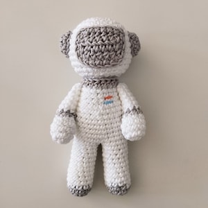 Pode incluir: Um brinquedo de astronauta de crochê branco com um capacete de crochê cinza e um pequeno remendo laranja e azul no peito.