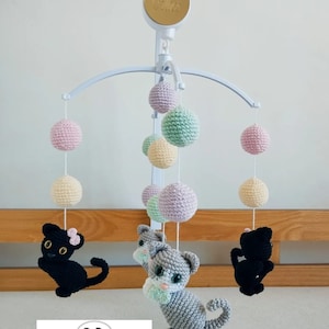 May include: A white baby mobile with crocheted gray and black kittens, pink, green, and yellow balls, and a gold musical box with the word "Alice" on it.