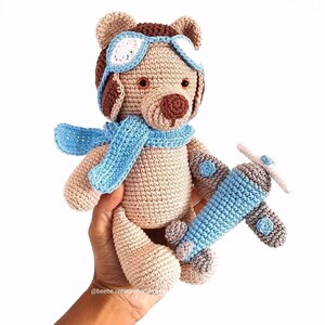 Pode incluir: Um urso de peluche bege feito em croché, usando um cachecol de aviador azul-claro e óculos a condizer com lentes brancas. O urso segura um pequeno avião de brinquedo em croché azul-claro com detalhes cinzentos. O texto @beebe.creativehandmade é visível.