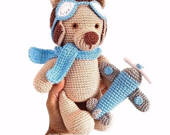 Puede incluir: Un oso de peluche beige tejido a ganchillo que lleva una bufanda de aviador azul claro y gafas a juego con lentes blancas. El oso sostiene un pequeño avión de juguete tejido en azul claro con detalles grises. El texto @beebe.creativehandmade es visible.