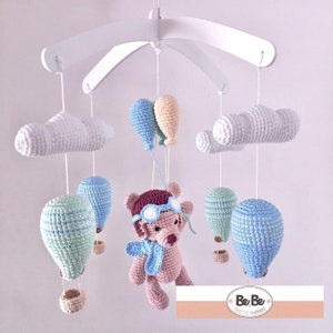 May include: A baby mobile with a crocheted teddy bear wearing aviator goggles and a blue scarf, surrounded by crocheted clouds and hot air balloons in shades of blue, green, and white. The mobile is suspended from a white wooden frame. The text "BeBe Creative Handmade" is visible in the lower right corner.