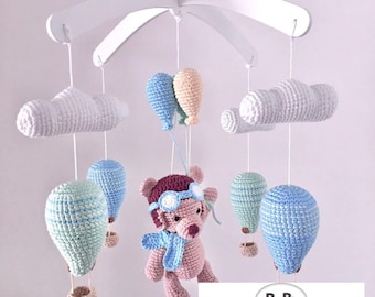 May include: A baby mobile with a crocheted teddy bear wearing aviator goggles and a blue scarf, surrounded by crocheted clouds and hot air balloons in shades of blue, green, and white. The mobile is suspended from a white wooden frame. The text "BeBe Creative Handmade" is visible in the lower right corner.