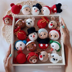 Op de afbeelding: Een verzameling handgemaakte gehaakte kerstversieringen in een witte doos. De ornamenten tonen verschillende feestelijke karakters, waaronder sneeuwmannen, kerstmannen, rendieren en beren. Elk ornament heeft een gouden ophanglus. Het kleurenpalet omvat rood, wit, bruin en zwart.