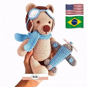 Peut inclure: Ours en peluche crocheté portant un chapeau d'aviateur marron avec des lunettes blanches et une écharpe bleue. L'ours tient un avion crocheté bleu et gris. L'ours est brun clair avec des accents brun foncé.