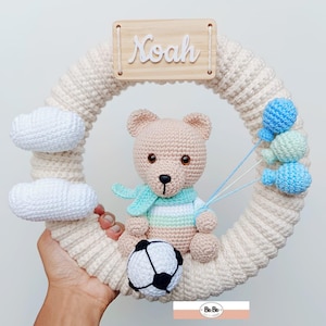 Pode incluir: Uma coroa de crochê com um urso de pelúcia marrom segurando uma bola de futebol. A coroa é decorada com nuvens brancas e balões azuis e verdes. Uma placa de madeira com o nome "Noah" está presa ao topo da coroa.