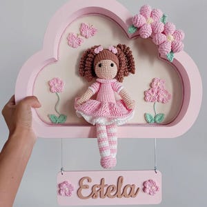 Pode incluir: Decoração de parede rosa em forma de nuvem com uma boneca de crochê feita à mão, cabelo castanho e vestido rosa. A moldura inclui flores de crochê, borboletas e uma placa com o nome "Estela".