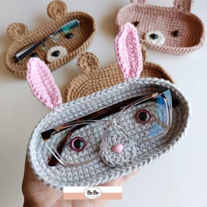 Pode incluir: Suportes de óculos de crochê em forma de animais em tons de castanho, rosa e cinza. O suporte cinza, em forma de coelho, contém óculos. Outros suportes têm formato de urso. Detalhes bordados e feitos à mão.