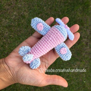 Pode incluir: Um pequeno avião de crochê com um corpo cilíndrico rosa, asas e cauda azul-claras e uma hélice branca. As asas são acentuadas com pequenos detalhes circulares cor-de-rosa. O texto beebe.creativehandmade é visível na parte inferior.