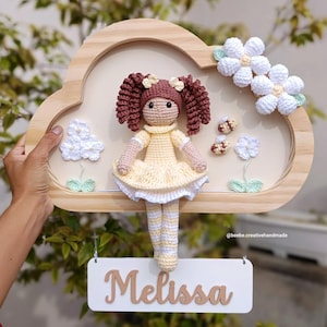 Pode incluir: Uma boneca de crochê feita à mão com cabelo castanho e um vestido amarelo é exibida dentro de uma moldura de madeira em forma de nuvem. A moldura é decorada com flores de crochê brancas e o nome "Melissa" em uma placa suspensa. A boneca tem cerca de 20 cm de altura.