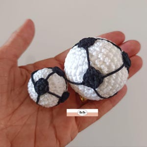 Pode incluir: São exibidas duas bolas de futebol de crochê, uma grande e outra pequena. As bolas são brancas com remendos pretos em forma de pentágono e costuras pretas. A bola menor tem aproximadamente 4 cm de diâmetro, enquanto a maior tem cerca de 6 cm de diâmetro.