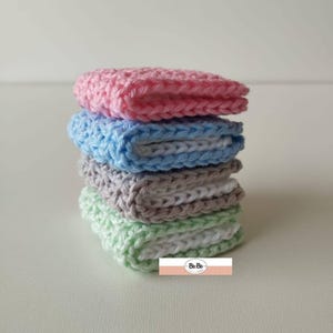 May include: A stack of four crocheted washcloths in pastel colors: pink, blue, gray, and green. The washcloths are neatly folded and stacked on top of each other. The bottom right corner of the image has a logo that says "Be Bo".