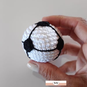 Puede incluir: Un pequeño balón de fútbol hecho a mano con ganchillo. El balón es blanco con formas de pentágono negro y costuras negras. El balón está en la mano de una persona.