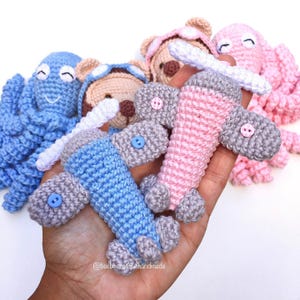 Puede incluir: Una colección de juguetes de ganchillo hechos a mano, que incluyen un pulpo azul, un pulpo rosa y varios aviones en azul y rosa. Los aviones tienen alas y hélices grises. Los juguetes se sostienen en una mano.