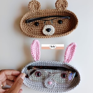 Pode incluir: Dois porta-óculos de crochê feitos à mão. O suporte superior é marrom e em forma de urso com olhos pretos e nariz branco. O suporte inferior é cinza e em forma de coelho com orelhas rosa e nariz rosa. Ambos os suportes têm óculos dentro.