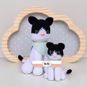 Pode incluir: Dois peluches de gato de crochê preto e branco com bandanas de padrão de estrelas verdes e brancas. O gato maior está sentado com o rabo enrolado em torno de suas pernas. O gato menor está sentado com o rabo atrás dele. Os gatos estão sentados na frente de uma moldura de madeira em forma de nuvem com um fundo cinza.