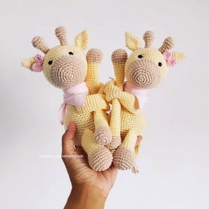 May include: Two handmade crochet giraffe toys in pale yellow with tan accents. Each giraffe features a light pink neck scarf, small pink flower details near the ears, and black bead eyes. The toys are crafted with a textured yarn stitch pattern.