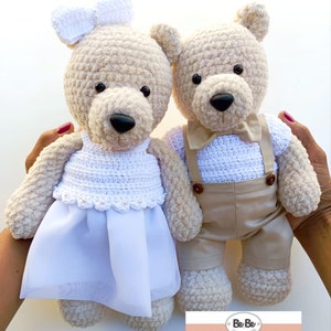 May include: Two handmade crochet teddy bears, one in a white dress with a bow and the other in a beige outfit with suspenders and a bow tie. The bears have black eyes and brown noses. The bears are held by hands.