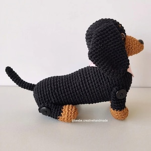 May include: A crocheted dachshund toy with a black body, tan paws, and a tan snout. The dog features black button joints at the legs, a light pink collar, and the text @beebe.creativehandmade printed near the bottom of the body.