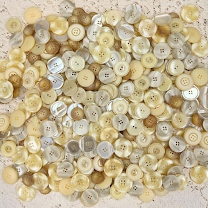 May include: A large collection of white and cream colored buttons of various sizes and styles. The buttons are made of a smooth, shiny material and have a variety of different designs.