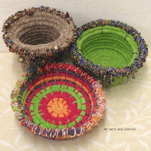 May include: Three handmade, colorful, beaded rope bowls. One is tan, cylindrical, and beaded with brown and gold beads. Another is green with blue and green beads. The third is a flat bowl with red, orange, and green concentric circles.